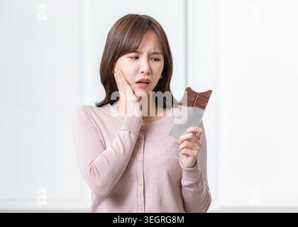 Une femme dans un pull rose tient une barre de chocolat, couvrant son visage d'une main, paraissant malheureuse dans une pièce blanche. Banque D'Images