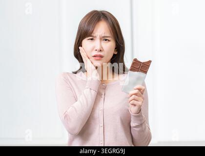 Une jeune femme tient une barre de chocolat et touche sa joue, paraissant inquiète dans une pièce blanche. Banque D'Images
