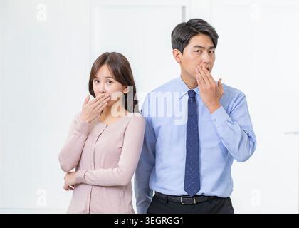 Une jeune femme et un jeune homme asiatiques se tiennent côte à côte, couvrant leur bouche dans une expression surprise, sur fond blanc. Banque D'Images