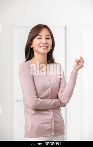 Une jeune femme asiatique se tient debout dans une pièce blanche lumineuse, souriant et faisant des gestes avec sa main. Banque D'Images