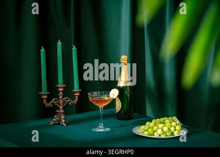 Une luxueuse bouteille de champagne, un verre à cocktail et une assiette de raisins sont posés sur une nappe de velours dans un cadre élégant et faiblement éclairé. Banque D'Images