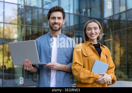 Jeunes collègues d'affaires debout à l'extérieur près d'un immeuble de bureaux en verre moderne, souriant à la caméra tout en partageant un ordinateur portable et une tablette, transmettant le travail d'équipe Banque D'Images
