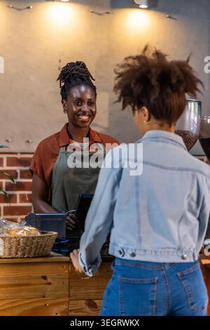 Barista afro-américaine dans un tablier vert qui exploite un point de vente tout en servant le client au comptoir du café Banque D'Images