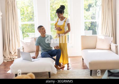 Couple diversifié enceinte dans des combinaisons jaunes berçant la bosse comme partenaire à l'aide d'un ordinateur portable sur pouf à la maison Banque D'Images