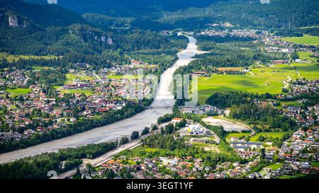 La vue aérienne montre la rivière qui coule à travers la ville de Reutte. Des maisons bordent les berges de la rivière, et des champs verdoyants répartis dans le paysage. Des arbres et des montagnes entourent la région. Banque D'Images