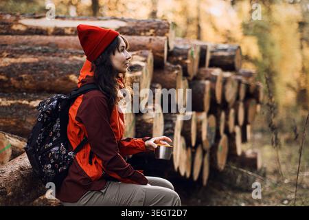 Femme s'assoit sur des bûches dans la forêt tout en tenant la tasse pendant la randonnée. Capacité à ralentir, améliorer la pleine conscience, réduire le stress, améliorer la prise de décision. Banque D'Images