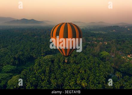 Vue aérienne d'une montgolfière rayée orange et noire flottant au-dessus d'une luxuriante forêt tropicale de palmiers et de collines brumeuses pendant l'heure dorée à Chandor, allez-y Banque D'Images