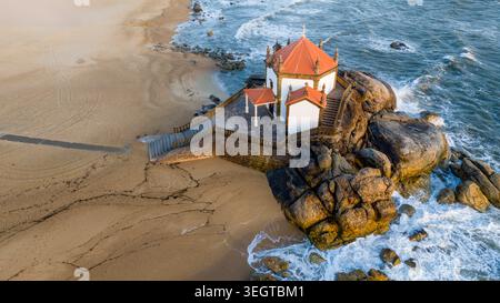 Vue aérienne de Capela do Senhor da Pedra sur un éperon rocheux entouré d'une plage de sable et de vagues écrasantes à Miramar, Porto, Portugal. Banque D'Images