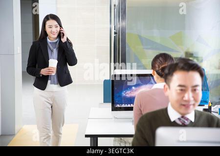 Une jeune femme asiatique en costume d'affaires entre dans un bureau moderne, parlant au téléphone tout en tenant une tasse de café. Banque D'Images