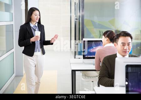 Une femme en costume d'affaires marche dans un bureau moderne, tenant une tasse à café et faisant des gestes à ses collègues à leur bureau. Banque D'Images