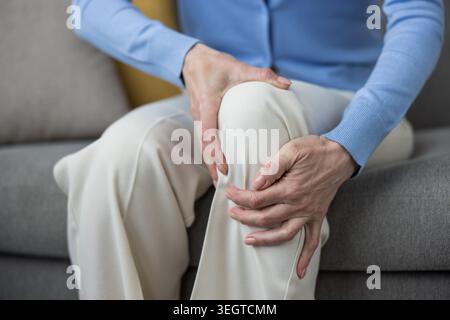 Femme mature assise sur un canapé, berçant son genou douloureux avec les deux mains, montrant l'inconfort de l'arthrite, des douleurs articulaires ou une blessure musculaire dans un cadre familial Banque D'Images