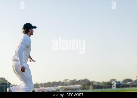 Mâle dans la vingtaine courir sur un terrain herbeux tenant une balle de cricket rouge portant un uniforme blanc, espace copie Banque D'Images