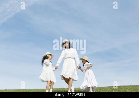 Une mère et ses deux jeunes filles marchent main dans la main à travers un champ verdoyant herbeux par une journée ensoleillée. Banque D'Images