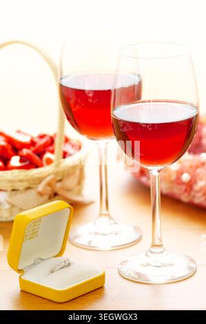 Une bague en diamant est présentée dans une boîte jaune à côté de deux verres de vin rouge sur une table en bois. Banque D'Images