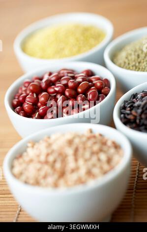 Une sélection de différents types de grains et de légumineuses, y compris les haricots rouges, le riz brun et le quinoa, sont disposés dans de petits bols blancs sur une surface en bois. Banque D'Images