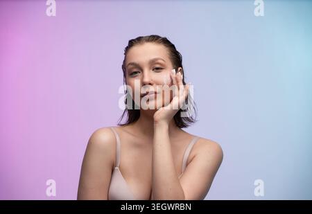 Jeune femme appliquant une crème pour le visage sur sa joue, favorisant une routine de beauté quotidienne pour une peau saine sur un fond coloré avec un espace de copie, en se concentrant sur Banque D'Images