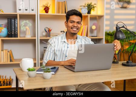 L'homme indien sur la table à la maison ouvre l'ordinateur portable avec un sourire joyeux et commence le travail à distance du matin. Arabian Guy sur la table à la maison commence la routine de bureau à domicile se sentant frais détendu et prêt pour les tâches en ligne à l'intérieur. Banque D'Images