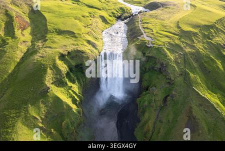 Vue aérienne de la cascade de Skogafoss descendant une falaise abrupte entourée de collines verdoyantes à Rangarthing eystra, Islande. Banque D'Images