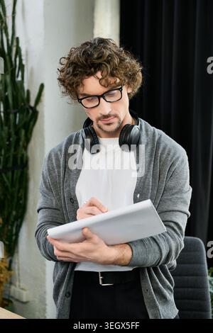 Un bel homme portant des lunettes et des écouteurs écrit des notes dans un espace de bureau élégant. Banque D'Images