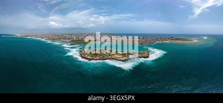 Vue aérienne de l'île de Ngor entourée de vagues turquoise avec la ligne d'horizon de la ville en arrière-plan à Dakar, région de Dakar, Sénégal. Banque D'Images