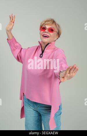 Une femme adulte, émotionnelle, espiègle, heureuse dans des vêtements roses et des lunettes avec ses mains levées Banque D'Images