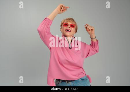 Une femme adulte, émotionnelle, espiègle, heureuse dans des vêtements roses et des lunettes avec ses mains levées Banque D'Images