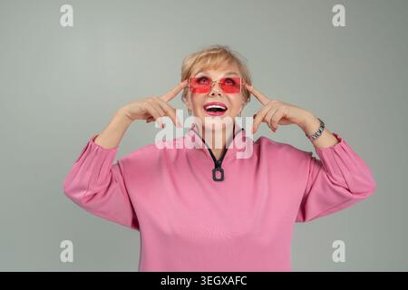 Une femme adulte, émotionnelle, espiègle, heureuse dans des vêtements roses et des lunettes avec ses mains levées Banque D'Images