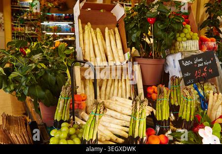 Asperges vertes et blanches fraîches disposées dans un marché avec des fruits et des plantes colorés visibles en arrière-plan. Banque D'Images