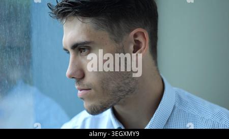 Portrait de jeune entrepreneur triste assis près de la fenêtre dans une chambre d'hôtel moderne ou un bureau. Homme malheureux avec une expression sérieuse regardant loin montrant Banque D'Images