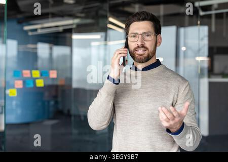 Jeune homme communiquant sur le téléphone portable, discutant du travail, debout dans l'intérieur de bureau contemporain, représentant la connexion, le professionnalisme et la communication à distance Banque D'Images