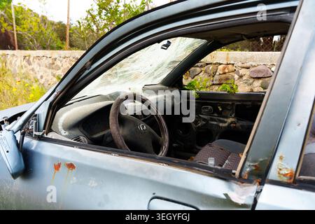 Vue intérieure d'un véhicule abandonné naufragé présentant un pare-brise brisé, une carrosserie rouillée et des panneaux de porte manquants dans un extérieur rural négligé Banque D'Images