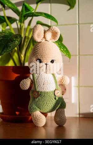 Lapin amigurumi crochet beige en salopettes en tricot vert et chemise blanche à côté d'une plante de feuilles en pot sur une surface en bois, jouet textile ensoleillé évoquant le charme fait main et l'émerveillement de l'enfance Banque D'Images