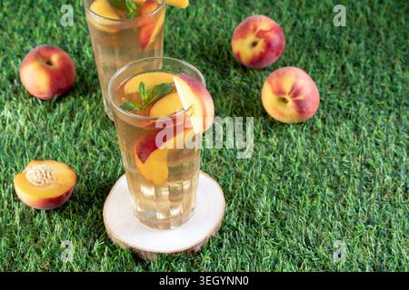 Des verres rafraîchissants de thé glacé à la pêche fait maison avec des feuilles de menthe et des fruits frais dispersés sur une pelouse verte vibrante Banque D'Images