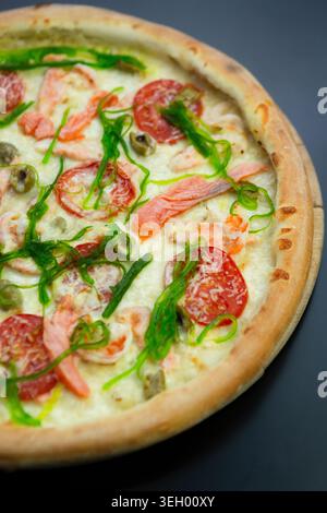 Une pizza est placée sur une planche de bois, surmontée de saumon, tomates, oignons verts et olives. La scène montre une atmosphère chaleureuse adaptée pour profiter de f Banque D'Images