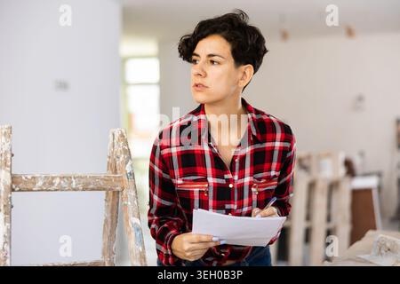 Portrait de jeune femme constructeur en chemise et short court à côté de l'escabeau avec des documents dans ses mains Banque D'Images