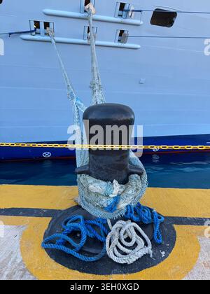 Une lourde borne d'amarrage noire avec des cordes enroulées autour d'elle sur un plancher de jetée peint en jaune et bleu contre un mur de bateau de croisière à Cabo Rojo. Banque D'Images
