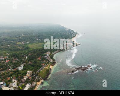 Vue aérienne de la côte tropicale avec des eaux turquoises, des affleurements rocheux et des palmiers verdoyants sous un ciel brumeux à Unawatuna, Provin du Sud Banque D'Images