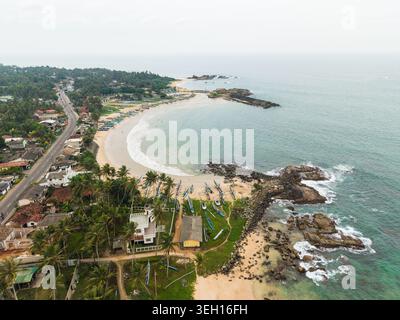 Vue aérienne d'une crique de plage de sable courbe avec des bateaux de pêche traditionnels sur le rivage et une route côtière Dodanduwa, Province du Sud, Sri Lanka. Banque D'Images