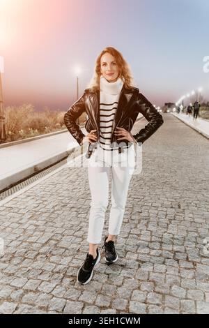 Femme Streetwear soirée femme élégante portant une veste en cuir et un Jean blanc pose sur une passerelle pavée au coucher du soleil Banque D'Images