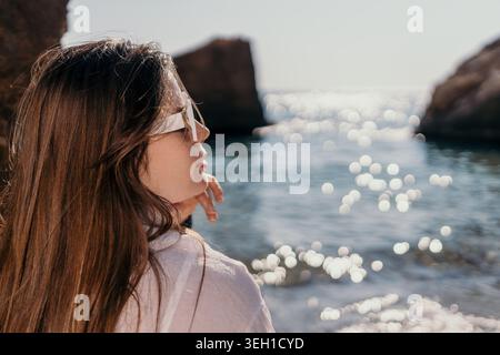 Femme coucher de soleil océan regardant les vagues scintillantes pendant l'heure dorée sereine tout en portant des lunettes de soleil blanches sur une plage rocheuse Banque D'Images