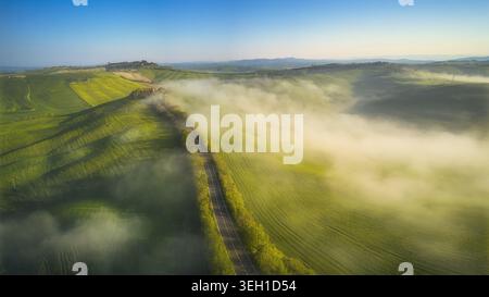 Vue aérienne des collines verdoyantes et une route sinueuse enveloppée de brume matinale sous un ciel bleu clair en Toscane, Italie. Banque D'Images
