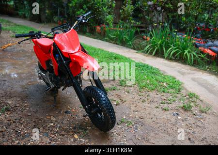 Moto de motocross rouge pour courses hors route garée sur un chemin de terre humide. puissant vélo de terre attend l'aventure et le sport extrême dans un environnement vert luxuriant extérieur c Banque D'Images
