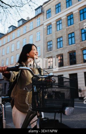 Jeune femme asiatique souriant tout en poussant son vélo dans une rue de la ville, portant des écouteurs et appréciant la musique. L'image transmet les déplacements urbains, happine Banque D'Images