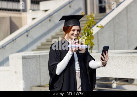 Femme portant une robe noire de diplômé et une casquette, debout sur la place du campus tenant le diplôme et le téléphone, souriant Banque D'Images