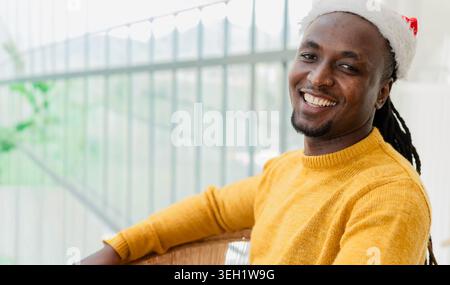 Homme afro-américain assis dans une chaise près des fenêtres, portant chapeau de Père Noël, pull moutarde, espace de copie Banque D'Images