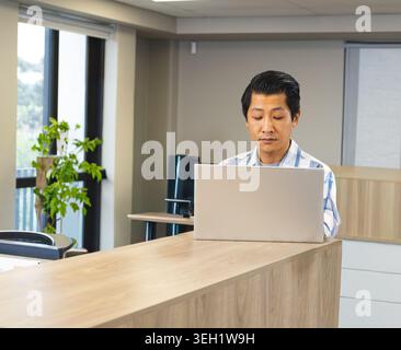 Homme asiatique mi-adulte portant chemise rayée travaillant à la réception avec ordinateur portable argenté et plante Banque D'Images