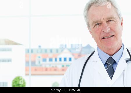 Médecin masculin principal debout à la fenêtre de la clinique portant une blouse blanche de laboratoire et un stéthoscope, espace de copie Banque D'Images
