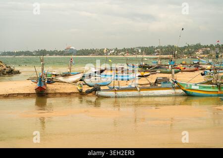 Vue des bateaux de pêche colorés traditionnels et des stabilisateurs reposant sur la côte sablonneuse avec une côte bordée de palmiers sous un ciel couvert Dodanduwa, donc Banque D'Images