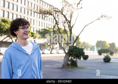 Adolescent masculin portant un sweat à capuche bleu clair souriant avec des lunettes rondes sur la rue urbaine, espace de copie Banque D'Images