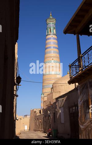 Islam Khoja Minaret vu à travers une ruelle étroite à Ichan Kala, Khiva. Des bandes de tuiles rayées et un dôme vert s'élèvent au-dessus des murs de briques de boue sous un bleu clair Banque D'Images